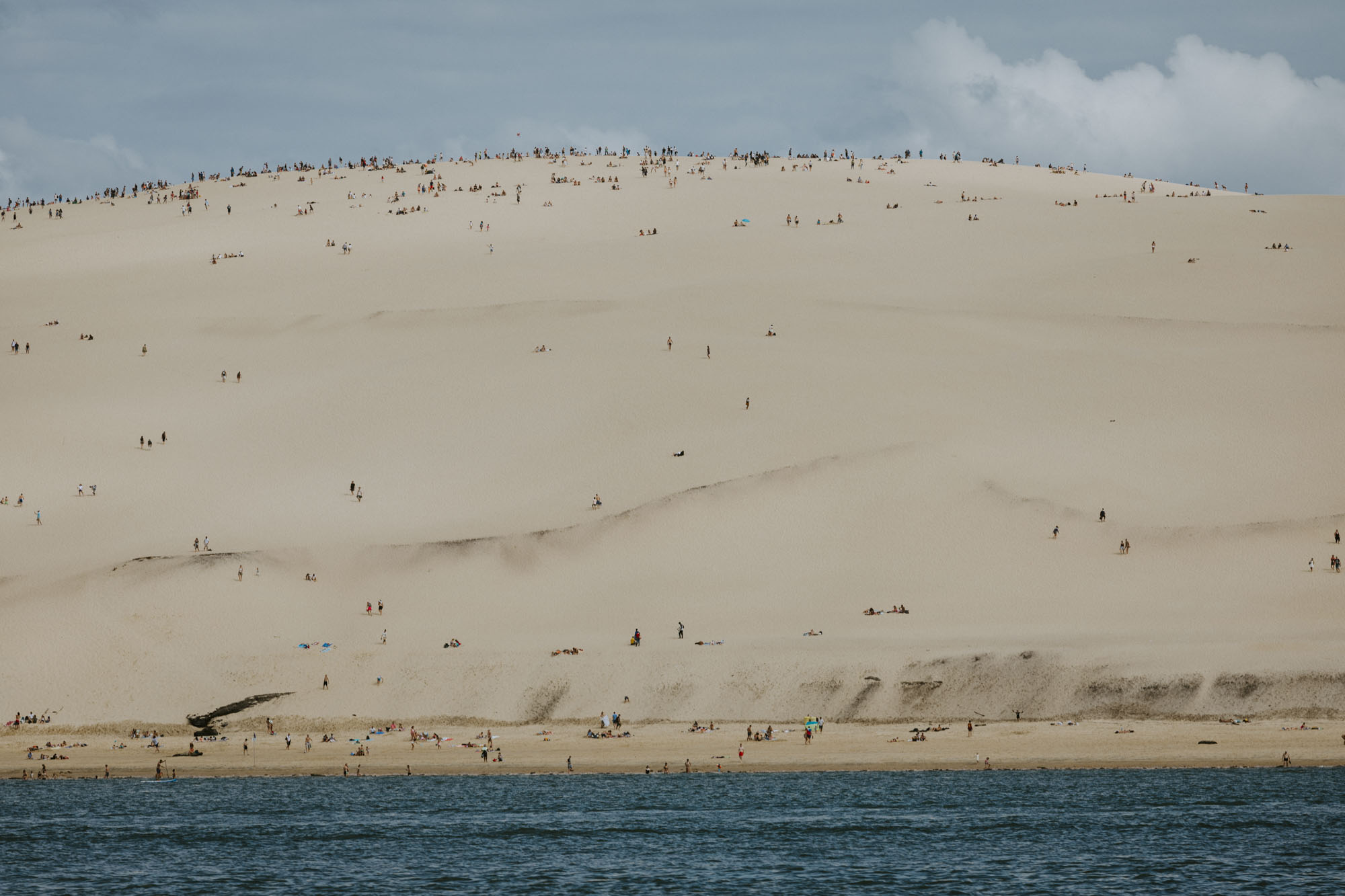 Arcachon wedding photographer: the bay boat is sailing at the botton of Dune du Pyla and all the wedding guests observe the tourists enjoying this landmark and looking like tiny dots